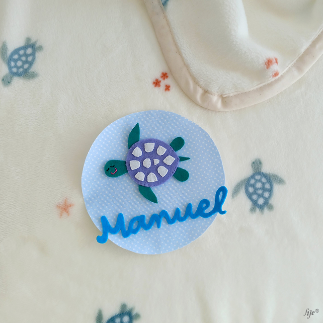 Etiqueta azul com tartaruga de feltro e nome Manuel sobre tecido branco com padrão de tartarugas