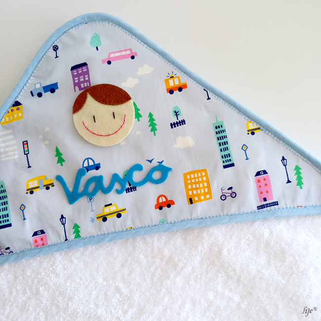 Toalha de bebé com capuz azul e padrões de carros e edifícios coloridos, com nome Vasco em feltro azul.