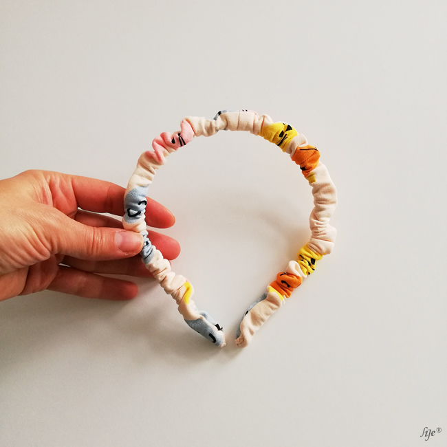 Tiara elástica franzida colorida sendo segurada numa mão sobre fundo branco.