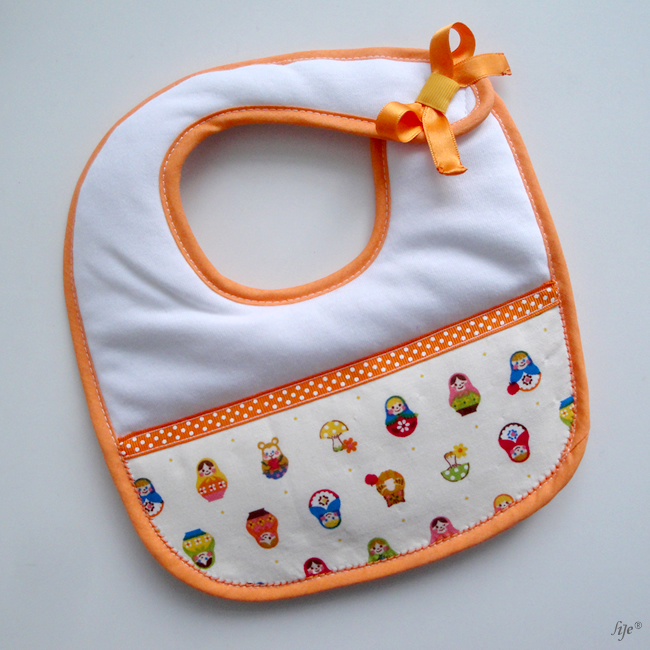 Babete branco com contorno laranja e padrão de bonecas russas coloridas