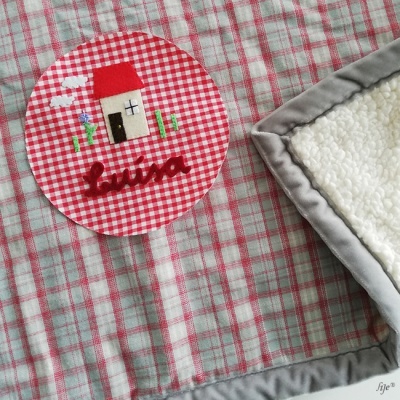 Detalhe de tecido xadrez vermelho e branco com bordado de casinha e nome 'luisa' junto a cobertor branco com bordas cinza