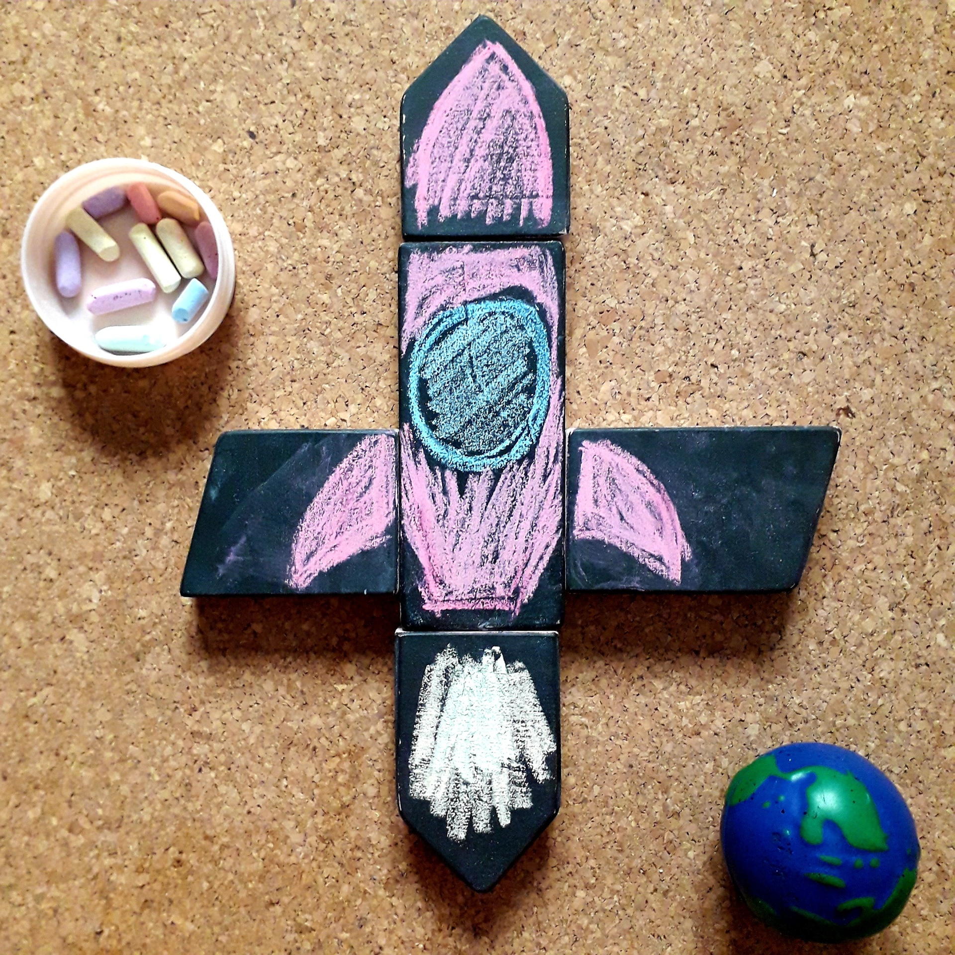 Cidade de Blocos Foguete de madeira com desenhos a giz, giz colorido numa taça e bola azul e verde sobre cortiça