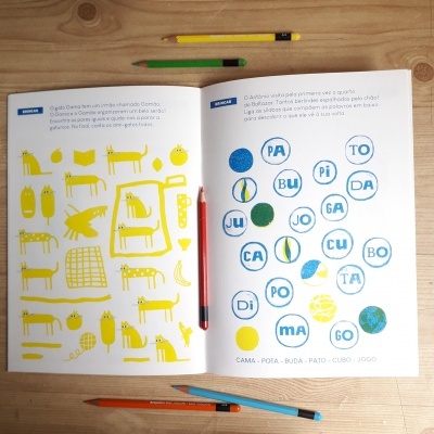 Livro aberto com desenhos amarelos e palavras dentro de círculos azuis, lápis coloridos à volta sobre mesa de madeira