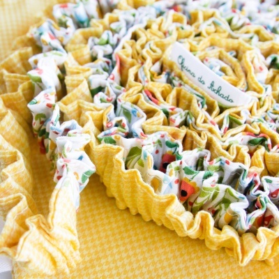 Detalhe de tecido amarelo xadrez e tecido branco com padrão floral, bordas franzidas e etiqueta bordada Casa da Abadá