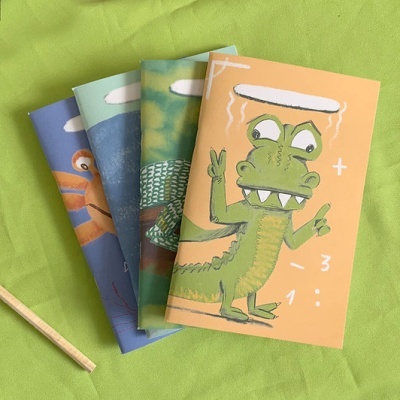 Quatro cadernos com capas ilustradas de animais em diferentes cores sobre tecido verde