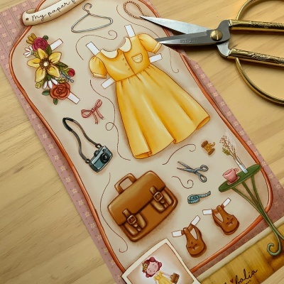 Página de papel recortável com desenhos de vestido amarelo, mala castanha, câmara preta, flores, animais e mesa verde sobre superfície de madeira clara com tesoura