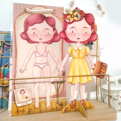 Boneca de papel vestida com vestido amarelo e mala castanha em suporte de madeira