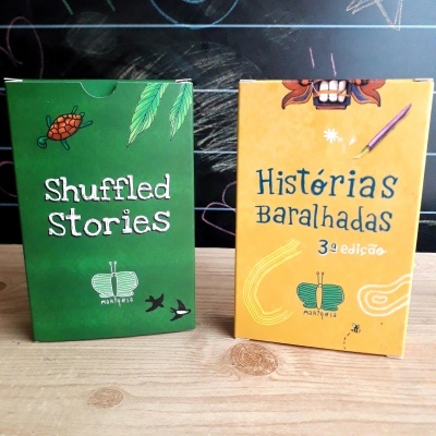 Dois baralhos de cartas, um verde e um amarelo, com textos 'Shuffled Stories' e 'Histórias Baralhadas 3ª edição'. 