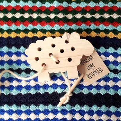 Brinquedo de madeira em forma de carneiro com cordão e etiqueta, sobre manta colorida em padrão de losangos.
