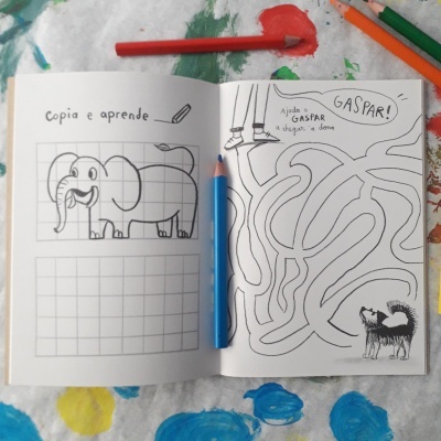 Livro infantil aberto com desenhos para copiar e labirinto, lápis de cor e fundo com tinta colorida