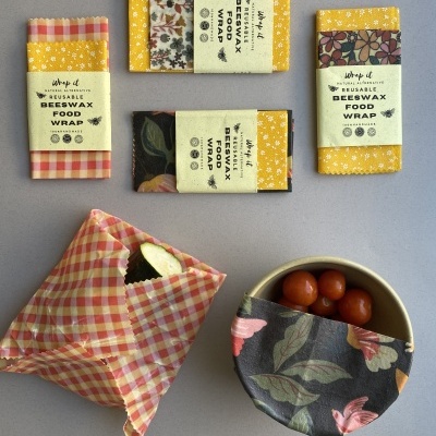 Quatro embalagens reutilizáveis de cera de abelha com diversos padrões coloridos, duas abertas com alimentos envoltos.