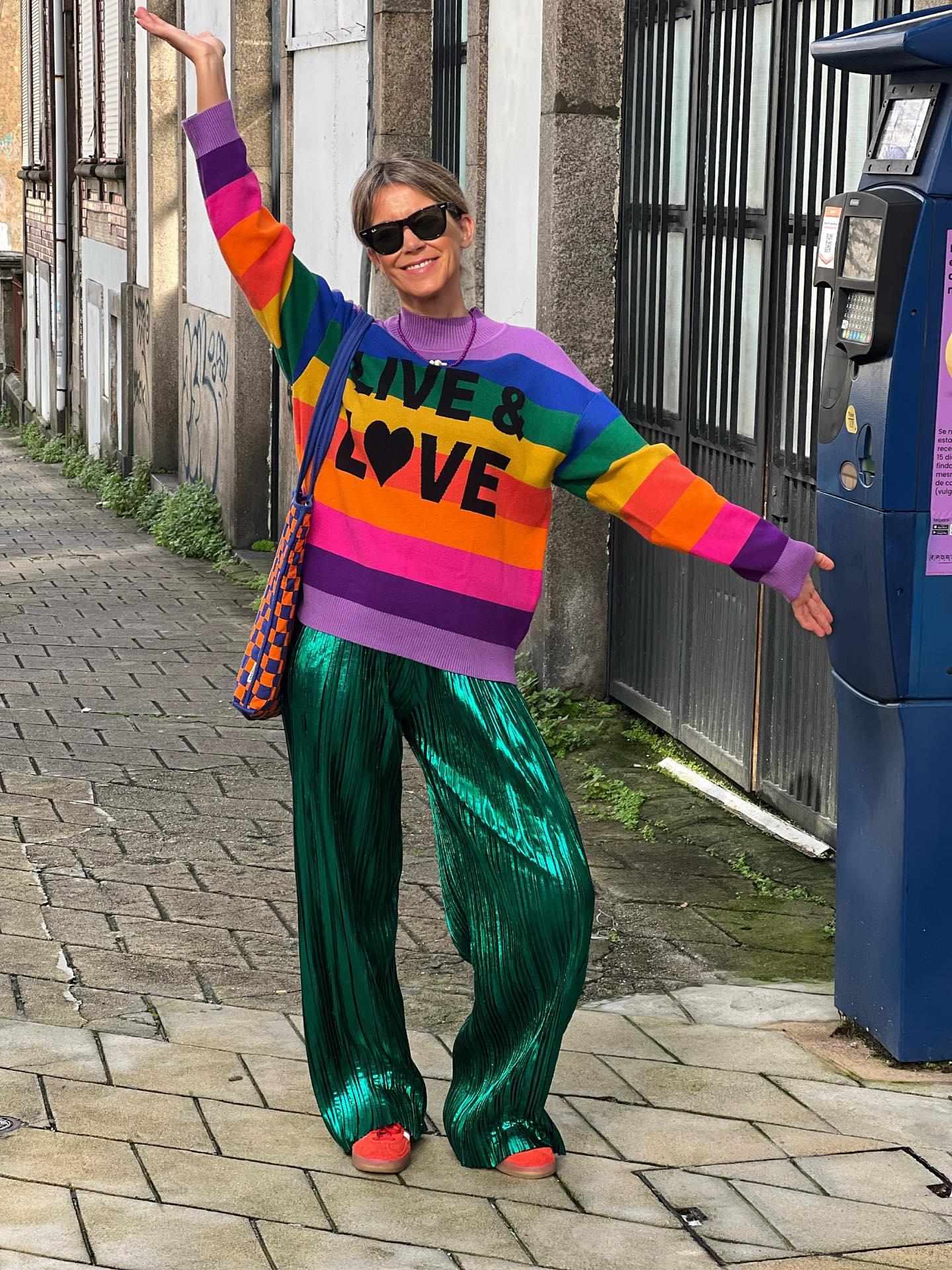 Mulher com camisola colorida com o texto LIVE & LOVE e calças verdes metalizadas na rua junto a máquina azul.