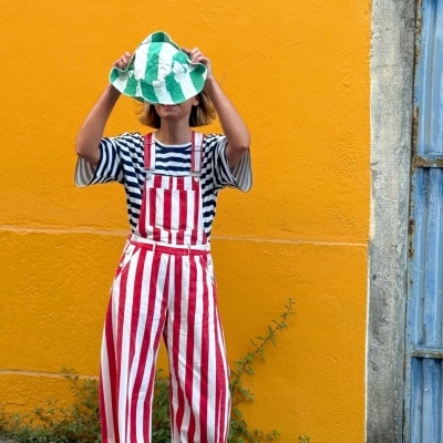 Pessoa em frente a parede laranja com jardineiras vermelhas e brancas, t-shirt às riscas azul e branco e chapéu verde e branco.