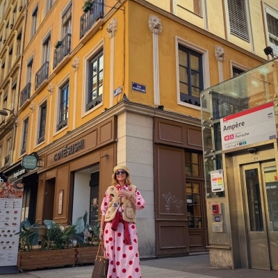 Mulher com vestido longo rosa de bolinhas vermelhas e colete bege, à frente de restaurante COTE SUSHI numa rua urbana.