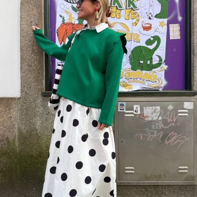 Mulher com vestido branco de bolinhas pretas, camisola verde de bordas onduladas e sapatos pretos em ambiente urbano.