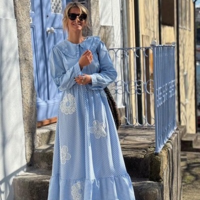 Mulher com vestido comprido azul claro às riscas e flores brancas, óculos de sol negros, em escadas junto a porta azul