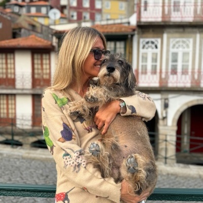 Mulher loira com camisola bege estampada a segurar cão cinza pelo longo em área urbana