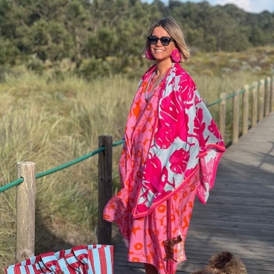 Mulher com vestido e xale florais, sapatos laranja, bolsa às riscas azul e vermelha, e cão junto a passadeira de madeira no exterior