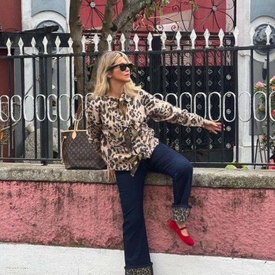 Mulher com casaco animal print e calças azuis com barra virada, sapatos vermelhos, óculos de sol e bolsa, junto a muro rosa e vedação metálica.