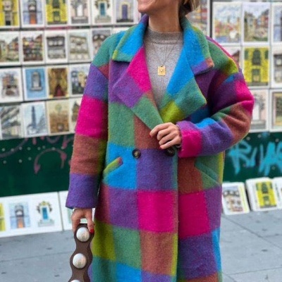 Casaco xadrez colorido felpudo usado por mulher com óculos e faixa amarela, segurando mala castanha com detalhes brancos, em ambiente urbano com cartazes.