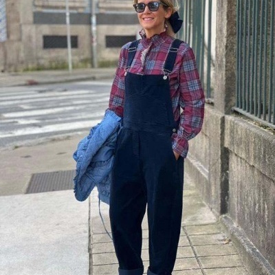 Mulher com camisa xadrez e jardineira preta numa calçada urbana