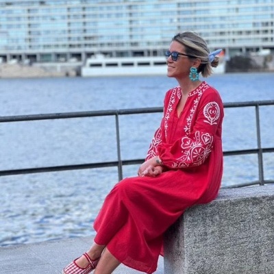 Mulher com vestido vermelho bordado sentada junto a um rio com óculos de sol e brincos azuis