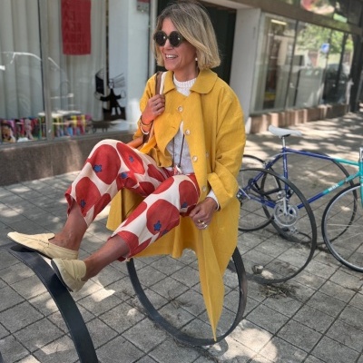 Mulher com casaco amarelo, calças padrão vermelho e branco, sapatos bege e óculos escuros sentada em suporte metálico junto a bicicleta