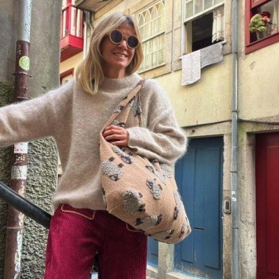 Mulher com suéter cinza, calças vermelhas e bolsa floral bege numa rua de pedra