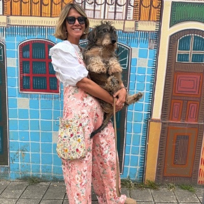 Mulher loira de óculos escuros com roupa floral rosa segurando cão contra mural colorido de paredes e portas.