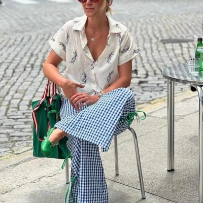 Mulher com calças xadrez azuis, blusa branca estampada, sapatos verdes e óculos vermelhos sentada numa esplanada urbana