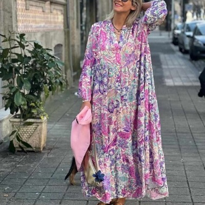 Mulher com vestido longo floral rosa e lilás, óculos escuros e sapatilhas douradas a caminhar na cidade