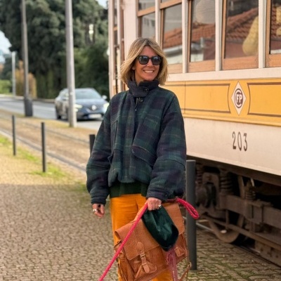 Mulher com casaco xadrez, calças laranja e mala castanha ao lado de elétrico amarelo.