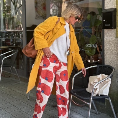 Mulher com casaco amarelo e calças com flores vermelhas junto a cadeira preta com mala bege numa esplanada