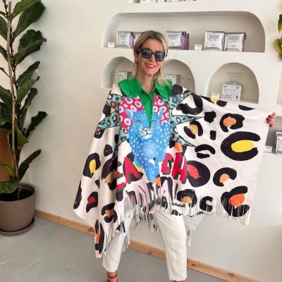 Mulher com poncho colorido em padrão de manchas e palavras, vestindo camisa verde e calças brancas, no interior com estantes e planta.