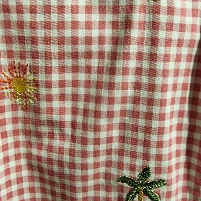 Tecido xadrez branco e rosa com bordados de flores e palmeiras e fita verde ondulada.