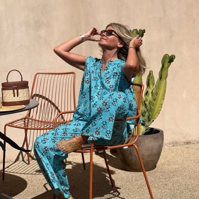 Mulher com roupa azul floral sentada numa cadeira laranja ao lado de planta e saco de palha