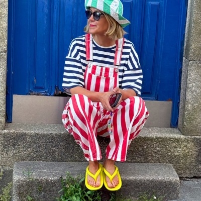 Pessoa sentada em degrau à frente de porta azul, vestindo roupa colorida com riscas e chapéu verde e branco