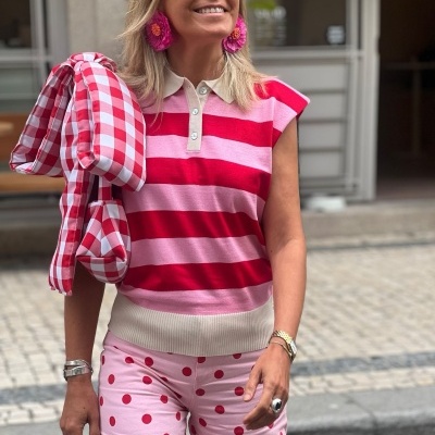 Mulher com colete riscado rosa e vermelho, calças rosa com bolinhas vermelhas, óculos de sol amarelos e brincos rosas