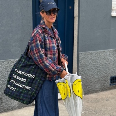 Mulher com casaco xadrez e boné segurando guarda-chuva transparente com carinhas amarelas e saco xadrez azul com texto em português