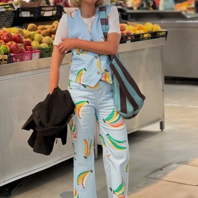 Conjunto azul claro com padrão de bananas, t-shirt branca, óculos escuros, sapatos castanhos e bolsa azul às riscas castanhas num mercado de frutas.