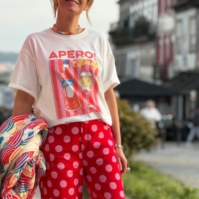 Pessoa com t-shirt branca estampada Aperol, calças vermelhas com bolinhas, óculos escuros e colar, segurando casaco colorido numa rua urbana