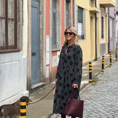 Mulher com vestido comprido cinzento de bolinhas pretas, mala vinho e sapatilhas pretas numa rua empedrada colorida