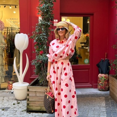 Mulher com conjunto de bolinhas vermelhas em fundo rosa, chapéu de palha e mala castanha em rua com parede vermelha
