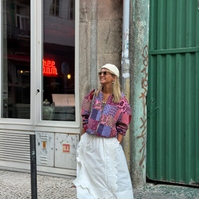 Mulher com casaco colorido e saia branca junto a parede e porta verde