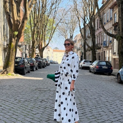 Mulher vestindo vestido branco com bolinhas pretas em rua de calçada com árvores e carros estacionados.