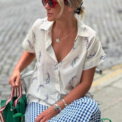 Mulher com camisa branca estampada, calças xadrez azul e branco, óculos de sol vermelhos e acessórios, sentada ao ar livre com bolsa verde.