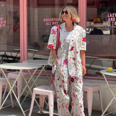 Mulher com fato floral branco e vermelho, mala vermelha, óculos escuros pretos, sapatos vermelhos, em esplanada com mesas e bancos rosas