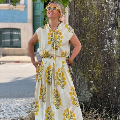 Vestido longo branco com flores amarelas e verdes, mulher com óculos amarelos, árvore ao fundo.
