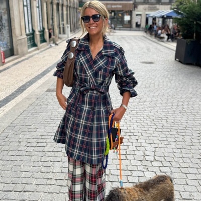 Mulher com casaco e calças às riscas num passeio urbano, segurando cão pela trela.