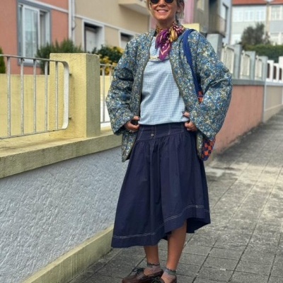 Mulher com casaco azul floral, saia azul, lenço roxo e óculos de sol amarelos numa calçada residencial